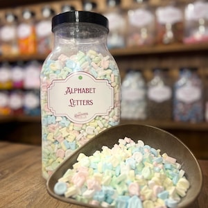 Könnte beinhalten: Ein klares Plastikglas gefüllt mit pastellfarbenen Buchstaben-Bonbons, beschriftet mit "Alphabet Letters". Eine kleine, braune Schale enthält einen Teil der Süßigkeiten. Die Süßigkeiten sind in Rosa, Blau, Gelb und Grün gehalten.