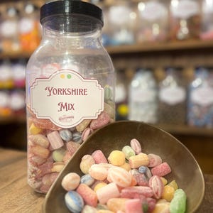 Pode incluir: Um frasco transparente de doces Yorkshire Mix com tampa preta, cheio de doces coloridos. Uma pequena concha de cor bronze contém uma porção dos doces. O rótulo no frasco diz "Yorkshire Mix". Os doces são de várias formas e cores.
