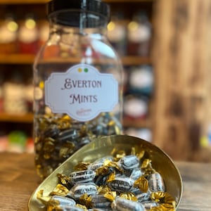 May include: A glass jar with a black lid and a label reading "Everton Mints" contains wrapped sweets. A brass dish holds a pile of the same sweets, some unwrapped. The sweets are dark with white stripes and wrapped in gold foil.