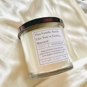 Puede incluir: Un tarro de vela de vidrio transparente con una tapa negra y una etiqueta blanca que dice "This Candle Smells Like You're Getting Married!" La etiqueta tambi&eacute;n incluye la fecha "29.08.24", la ubicaci&oacute;n "INN ON THE LAKE" y un mensaje que dice "Happy Birthday to the Bride! We cannot wait for your big day, it's going to be magical. All our love xoxo". La etiqueta est&aacute; firmada "Midnight Bride" e incluye el tiempo de combusti&oacute;n "40 hrs".
