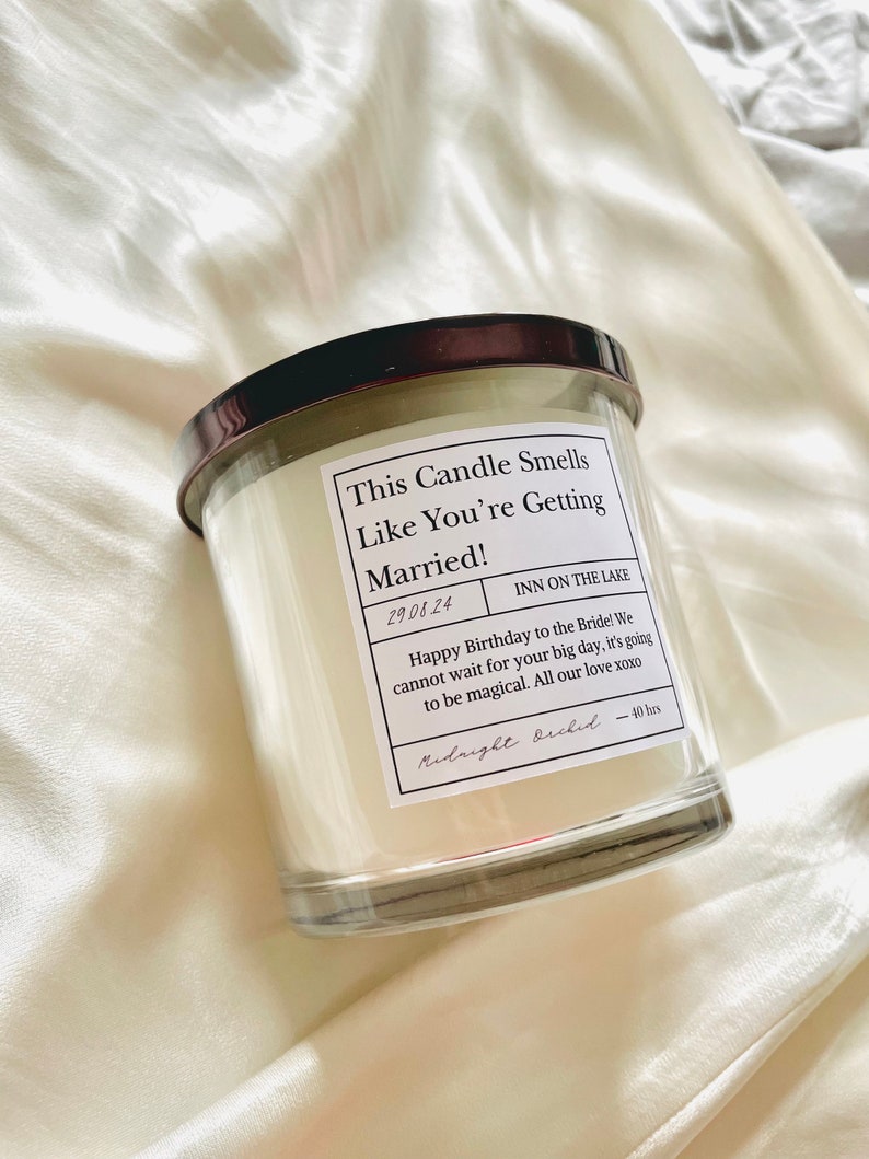 Puede incluir: Un tarro de vela de vidrio transparente con una tapa negra y una etiqueta blanca que dice "This Candle Smells Like You're Getting Married!" La etiqueta tambi&eacute;n incluye la fecha "29.08.24", la ubicaci&oacute;n "INN ON THE LAKE" y un mensaje que dice "Happy Birthday to the Bride! We cannot wait for your big day, it's going to be magical. All our love xoxo". La etiqueta est&aacute; firmada "Midnight Bride" e incluye el tiempo de combusti&oacute;n "40 hrs".
