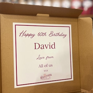 May include: A brown cardboard box with a white card inside. The card has a burgundy border and the text "Happy 60th Birthday David, Love from, All of us" and a confectioners logo.