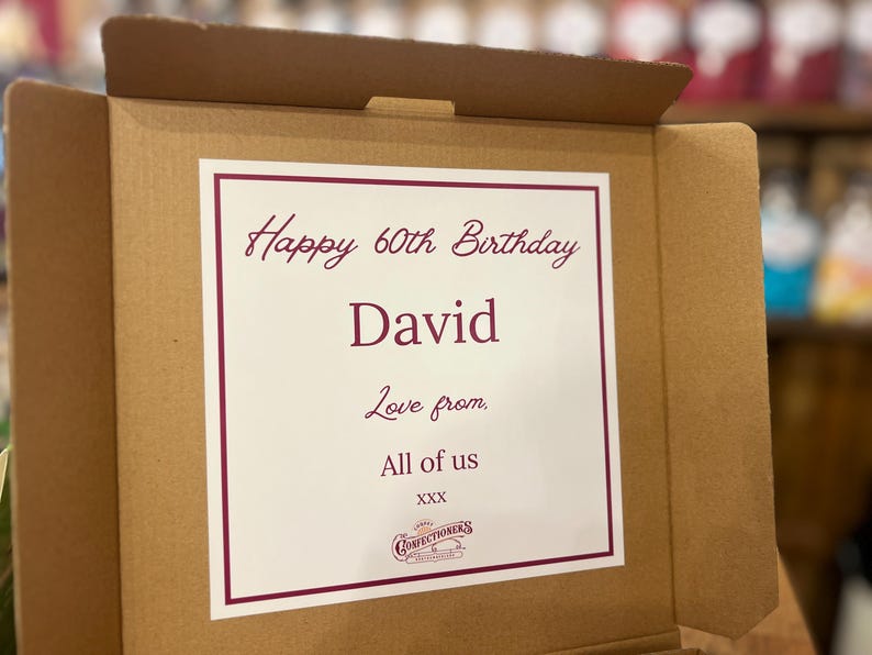 May include: A brown cardboard box with a white card inside. The card has a burgundy border and the text "Happy 60th Birthday David, Love from, All of us" and a confectioners logo.