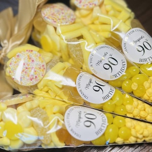 May include: A selection of clear cellophane bags filled with yellow sweets, tied with a gold ribbon. Each bag features a sticker saying "Thank you for coming to Margaret's 90th Birthday!" The bags contain jelly beans and other treats.