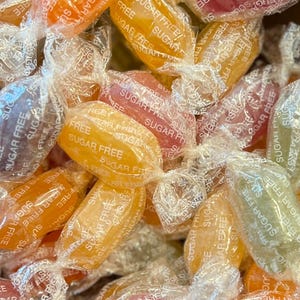 Könnte beinhalten: Nahaufnahme eines Haufens farbenfroher, einzeln verpackter Bonbons. Die Bonbons sind in den Farbtönen Orange, Gelb, Lila und Grün gehalten. Jeder transparente Wrapper ist mit dem Wort "SUGAR FREE" bedruckt.