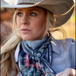 Op de afbeelding: Een vrouw draagt een lichtgekleurde cowboyhoed met een patroon op de kroon en rand. Ze draagt ook een blauwe sjaal met een bloemen- en geometrisch ontwerp en een lichtblauw overhemd.
