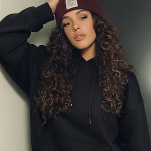 Può includere: Un berretto bordeaux con un'etichetta quadrata bianca con la scritta "SHARP FIGHT" è indossato con una felpa con cappuccio nera. La modella ha lunghi capelli castani ricci e si appoggia a un muro.