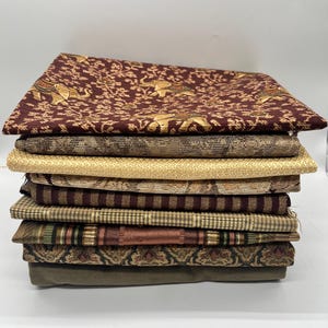 May include: A stack of various fabric samples in shades of brown, tan, and olive green. The top fabric is a deep burgundy with gold elephant and floral patterns. Other fabrics include striped, textured, and patterned designs.