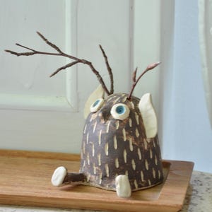 Könnte beinhalten: Eine skurrile Eulenfigur aus Keramik. Der braune Körper hat weiße Akzente und blaue Augen. Sie hat zweigartige Details und sitzt auf einem Holztablett. Ein einzigartiger Dekorationsartikel, der Charme in jedes Zuhause bringt.