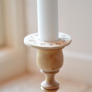 May include: A wooden candlestick with a white candle. The base of the candlestick has a ring with the letters "DECEMBER" engraved on it.