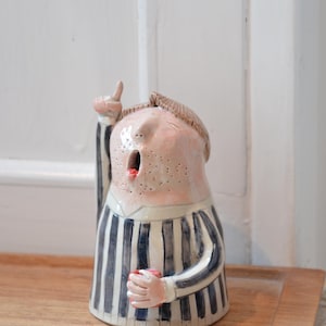 Może przedstawiać: Ceramiczna figurka osoby z zaskoczoną miną, wskazującej w górę. Postać ma różową twarz z ciemnymi piegami, krótkie brązowe włosy i ubrana jest w niebiesko-biały pasiasty strój. Trzyma mały czerwony przedmiot.