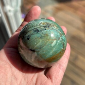 Puede incluir: Una esfera pulida de serpentina, con tonos de verde, marr&oacute;n y blanco. La esfera tiene una superficie lisa y brillante y es aproximadamente del tama&ntilde;o de una pelota de b&eacute;isbol.