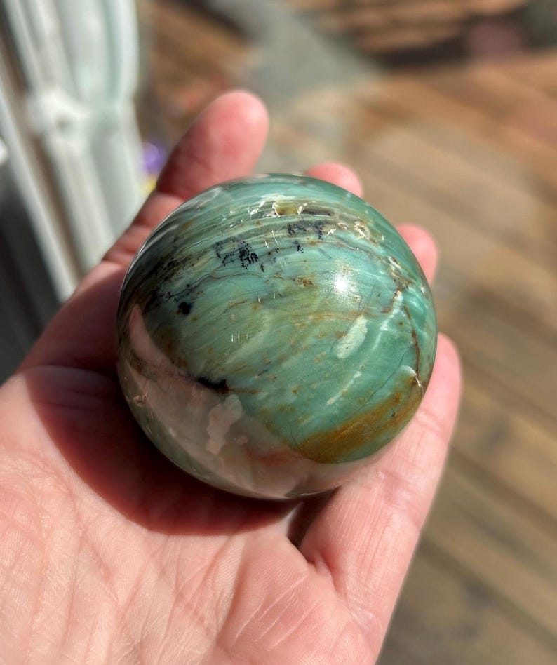 Puede incluir: Una esfera pulida de serpentina, con tonos de verde, marr&oacute;n y blanco. La esfera tiene una superficie lisa y brillante y es aproximadamente del tama&ntilde;o de una pelota de b&eacute;isbol.