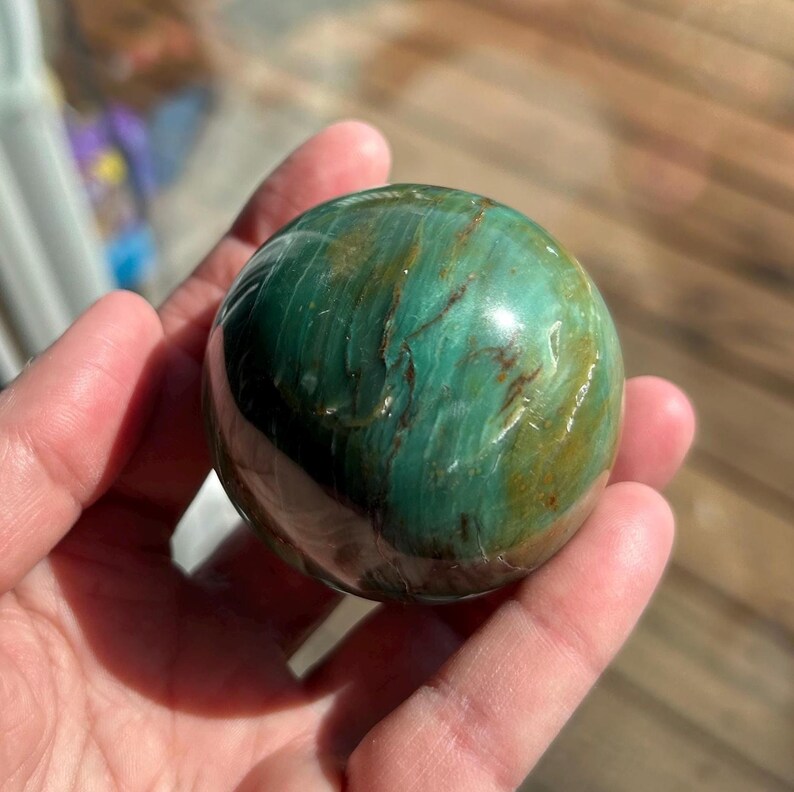 Puede incluir: Una esfera de piedra serpentina pulida de color verde y marr&oacute;n. La esfera tiene un patr&oacute;n de remolino de verde y marr&oacute;n.