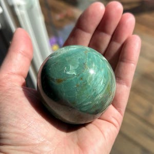 Puede incluir: Una esfera verde y marr&oacute;n de piedra serpentina pulida. La esfera tiene un acabado liso y brillante con intrincados patrones de verde y marr&oacute;n que se arremolinan.