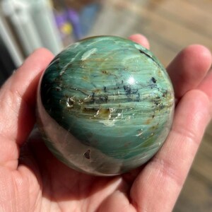 Puede incluir: Una esfera verde y marr&oacute;n con un patr&oacute;n de remolino, pulida a un acabado liso. La esfera est&aacute; hecha de una piedra natural, posiblemente &oacute;palo o jaspe.