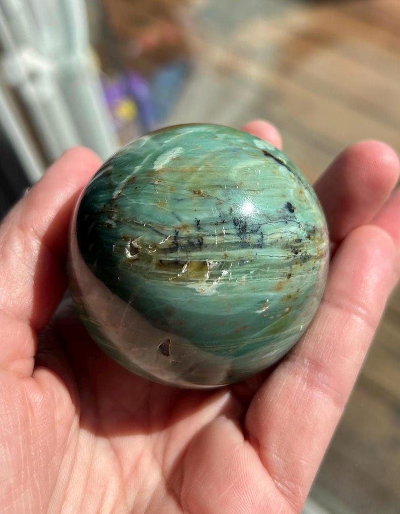 Puede incluir: Una esfera verde y marr&oacute;n con un patr&oacute;n de remolino, pulida a un acabado liso. La esfera est&aacute; hecha de una piedra natural, posiblemente &oacute;palo o jaspe.