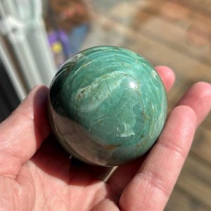 Puede incluir: Una esfera de piedra serpentina pulida de color verde y marr&oacute;n. La esfera es lisa y tiene un acabado brillante.