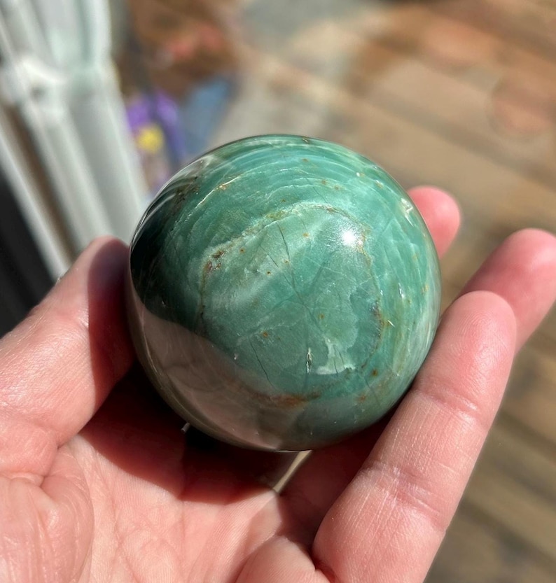 Puede incluir: Una esfera de piedra serpentina pulida de color verde y marr&oacute;n. La esfera es lisa y tiene un acabado brillante.