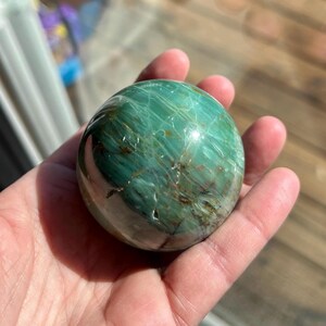 Puede incluir: Una esfera verde y marr&oacute;n de serpentina pulida, un tipo de piedra preciosa, se sostiene en una mano. La esfera tiene una superficie lisa y pulida y un patr&oacute;n de remolino de verde y marr&oacute;n.