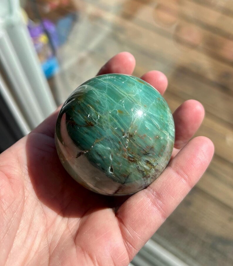 Puede incluir: Una esfera verde y marr&oacute;n de serpentina pulida, un tipo de piedra preciosa, se sostiene en una mano. La esfera tiene una superficie lisa y pulida y un patr&oacute;n de remolino de verde y marr&oacute;n.
