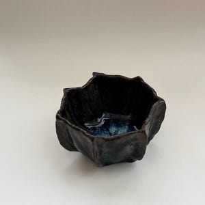 Peut inclure: Un petit bol en céramique brun foncé avec un glaçage bleu et vert à l'intérieur. Le bol a une forme unique et irrégulière avec de nombreux points.