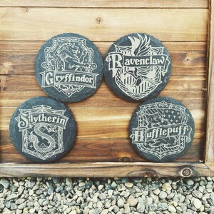 May include: Four black slate coasters with white designs of the Hogwarts house crests. The crests are Gryffindor, Ravenclaw, Slytherin, and Hufflepuff.