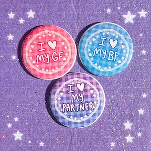 May include: Three circular pin-back buttons featuring a gingham pattern and a white lace-style border. The pink button reads I LOVE MY GF, the blue button reads I LOVE MY BF, and the purple button reads I LOVE MY PARTNER, each with a white heart.