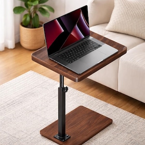 May include: A dark brown, adjustable laptop table with a black metal base. A silver laptop is placed on the table. The table is positioned in front of a white couch and a potted plant.