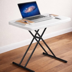 May include: A white, rectangular folding table with black metal legs. A laptop, notebook, smartwatch, mouse, and phone are on the table. The laptop screen displays a galaxy image. The table is on a hardwood floor.
