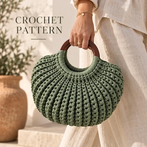 Puede incluir: Un bolso de ganchillo color verde salvia con una textura acanalada redondeada y dos asas circulares de madera marrón oscuro. El texto CROCHET PATTERN está impreso en gris oscuro sobre el bolso, sostenido por una mano con pulsera y anillo de oro.