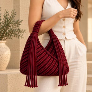 May include: A deep burgundy handbag crafted from thick cord in a macrame style. The bag features a triangular silhouette with a structured handle and long, dangling fringe accents that extend from the top attachment points downward.