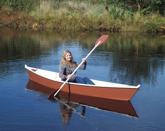 May include: A woman paddles a small, wooden canoe on a calm lake. The canoe is painted red and white, and the woman is wearing a blue shirt and jeans.