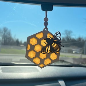 May include: A hexagonal wooden car air freshener featuring a honeycomb pattern with translucent yellow inserts and a black bee silhouette attached to the front. The ornament hangs from a string with a small wooden bead, suspended from a rearview mirror.