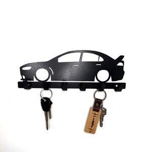 May include: Black metal key holder shaped like a car silhouette with five hooks. The key holder has a wooden keychain with the text "Life is better with a car" on it.