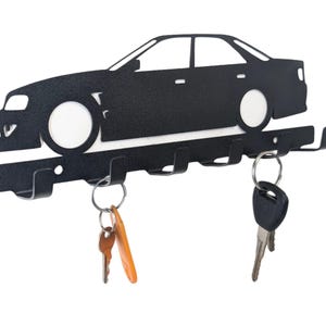 Pode incluir: Porta-chaves em metal preto em forma de carro com cinco ganchos. A silhueta do carro tem círculos brancos para as rodas e segura chaves. Um item elegante e funcional para os entusiastas de carros.