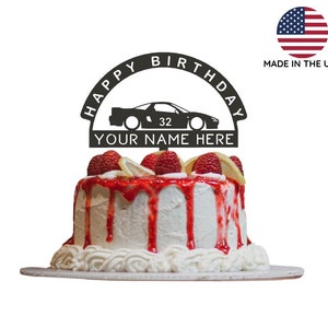 Puede incluir: Topper de pastel de metal negro con las palabras "Happy Birthday" en forma de arco, una silueta de un coche y el texto "Your Name Here". El topper está en un pastel blanco con glaseado rojo y fresas.