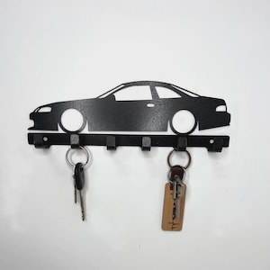 以下が含まれることがあります： 黒い金属製の車の形をしたキーホルダー。キーホルダーには、車のシルエットの下に5つのフックがあります。2組の鍵がフックからぶら下がっています。キーホルダーは白い壁に取り付けられています。
