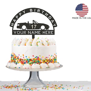 May include: Black cake topper featuring a silhouette of a convertible car with the words "Happy Birthday" and "Your Name Here". The number 17 is visible on the car. The topper is on a white cake with colourful sprinkles.