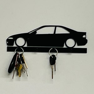 May include: Black metal key holder in the shape of a car. The key holder has a horizontal bar with key hooks and a car silhouette above. Two sets of keys hang from the hooks.