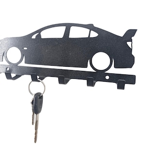 May include: A black metal car-shaped key holder with five hooks. The key holder is a silhouette of a car with a spoiler. A set of keys hangs from one of the hooks. The key holder is mounted on a white background.