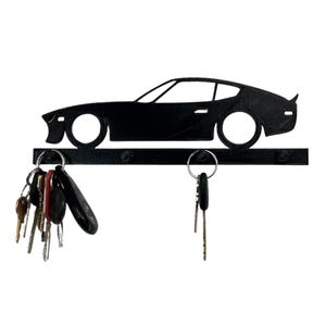 Puede incluir: Llavero de metal negro con diseño de silueta de coche. El llavero tiene una barra horizontal con llaveros y un contorno de coche encima. Varias llaves cuelgan de los anillos. Un artículo de decoración del hogar elegante y funcional.