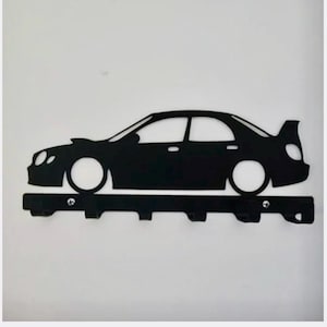 May include: A black metal wall-mounted key holder featuring a silhouette of a saloon car with a rear spoiler. The horizontal base bar includes six downward-facing hooks for hanging items and is attached with two visible silver-coloured screws.