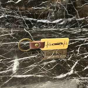 Könnte beinhalten: Ein Holzschlüsselanhänger mit einem braunen Lederband und einem Metallring. Der Schlüsselanhänger zeigt ein stilisiertes Auto mit einem Herzschlag-Motiv.