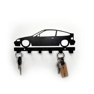 Puede incluir: Llavero de metal negro con forma de coche, con cinco ganchos. La silueta del coche tiene ruedas circulares grandes y un diseño elegante. Dos juegos de llaves cuelgan de los ganchos.