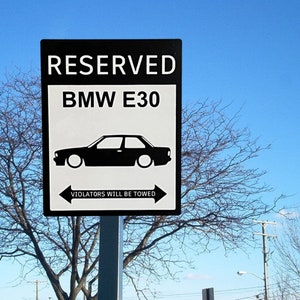 May include: A black and white sign with the text "RESERVED BMW E30" and a car silhouette. Below the car is the text "VIOLATORS WILL BE TOWED" with arrows pointing left and right. The sign is mounted on a metal pole against a blue sky.