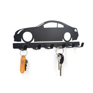 May include: A matte black metal wall-mounted key holder featuring a silhouette of a sports car. The horizontal base includes six upward-curving hooks for hanging keys. Two sets of keys are shown hanging from the hooks, one with an orange tag.