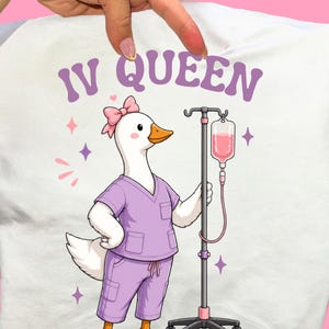Könnte beinhalten: Weißes T-Shirt mit einer Cartoon-Gans in lila OP-Kleidung und einer rosa Schleife, die einen Infusionsständer mit einem rosa Flüssigkeitsbeutel hält. Der Text "IV QUEEN" steht in Lila über der Gans, mit rosa und lila Sternakzenten.
