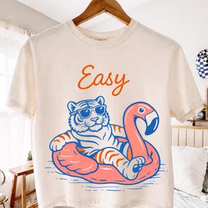 Könnte beinhalten: Cremefarbenes T-Shirt mit einem Tiger mit Sonnenbrille auf einem rosa Flamingo-Schwimmring. Der Schriftzug "Easy" ist in Orange gehalten. Das T-Shirt hängt an einem Holzkleiderbügel.