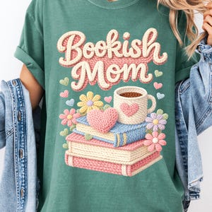 May include: Green t-shirt with the text "Bookish Mom" in a knitted style. The design includes a stack of books, a coffee mug, flowers, and hearts. The shirt has rolled-up sleeves and is worn with a denim jacket.